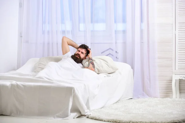 Headache concept. Guy on painful face waking up in morning. Man laying on bed, touching head , white curtain on background. Macho with beard and mustache suffers from headache, hangover in morning — Stock Photo, Image