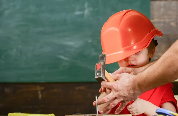 Παιδί αγόρι σφυρηλάτηση των καρφιών σε ξύλινη σανίδα. Νήπιο στο πολυάσχολο πρόσωπο παίζει στο σπίτι σε εργαστήριο. Νέα αντίληψη δεξιότητες. Το παιδί με πολύ μεγάλο κράνος χαριτωμένο μαθαίνοντας πώς να καρφιά σφυρί με αρσενικό τα χέρια βοηθείας — Φωτογραφία Αρχείου