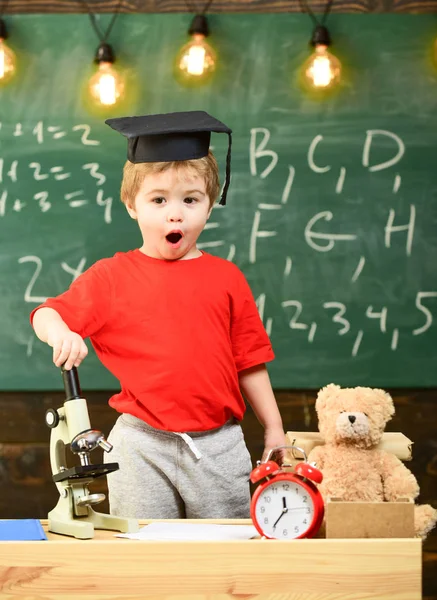 Niño, pupila en la cara conmocionada cerca del microscopio. Niño con gorra de graduado cerca del microscopio en el aula, pizarra en el fondo. Primero interesado en estudiar, educación. Concepto Wunderkind — Foto de Stock