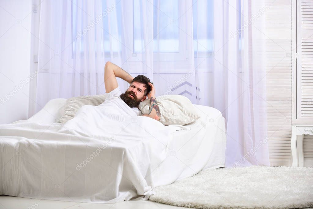 Headache concept. Guy on painful face waking up in morning. Man laying on bed, touching head , white curtain on background. Macho with beard and mustache suffers from headache, hangover in morning