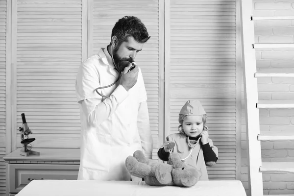 Salud y concepto de infancia. Hombre con barba y niño hablar en estetoscopio sobre fondo de madera. El veterinario y el ayudante examinan al oso de peluche. Padre e hijo — Foto de Stock