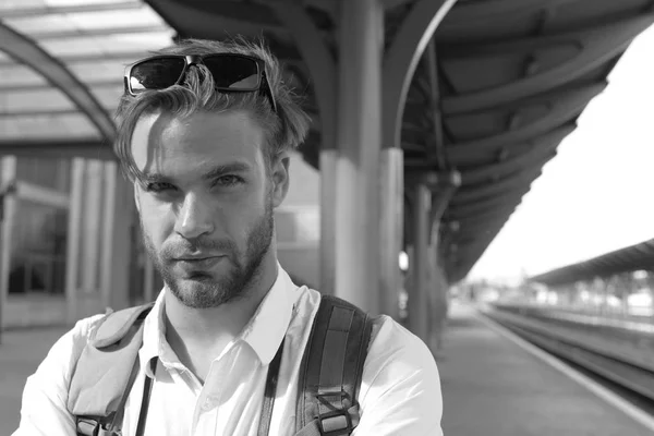 Guy with sunglasses waits for train, defocused. Missed train and travelling concept. Tourist with serious faceand backpack. Young man standing on platform at train station, close up.
