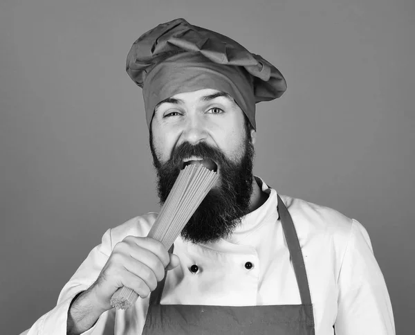 Concept de cuisine italienne. Homme ou hipster barbu tient des macaronis sur fond bleu. Chef avec des spaghettis. Cuisiner avec le visage affamé — Photo