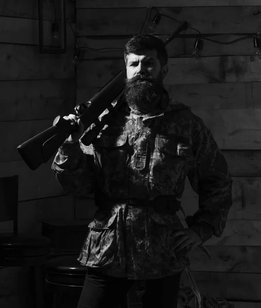 Gamekeeper concept. Hunter, brutal hipster on strict face with gun ready for hunting. Man with beard wears camouflage clothing, carries rifle on shoulder, wooden interior background