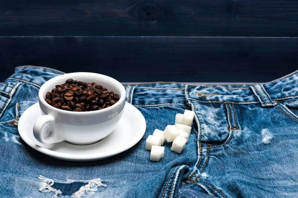 Kopp med kaffe bönor, raffinerat socker låg på jeans nära zip, denim bakgrund. Nybryggt kaffe konceptet. Mugg full av kaffebönor på jeans — Stockfoto