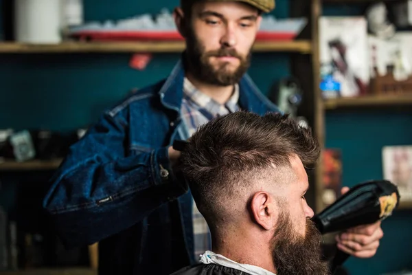 Hipster bearded client getting hairstyle. Styling concept. Barber with hairdryer drying and styling hair of client. Barber with hairdryer works on hairstyle for bearded man, barbershop background