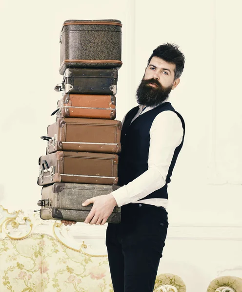 Butler and service concept. Man with beard and mustache wearing classic suit delivers luggage, luxury white interior background. Macho, elegant porter carries heavy pile of vintage suitcases