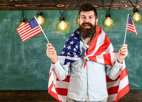 Conceito de educação patriótica. Professora americana acena com bandeiras americanas. Programa de intercâmbio de estudantes. Homem com barba e bigode no rosto feliz segura bandeiras dos EUA, em sala de aula, quadro-negro no fundo — Fotografia de Stock