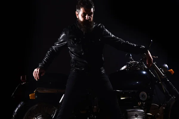 Macho, brutal biker in leather jacket stand near motorcycle at night time, copy space. Man with beard, biker in leather jacket lean on motor bike in darkness, black background. Biker culture concept
