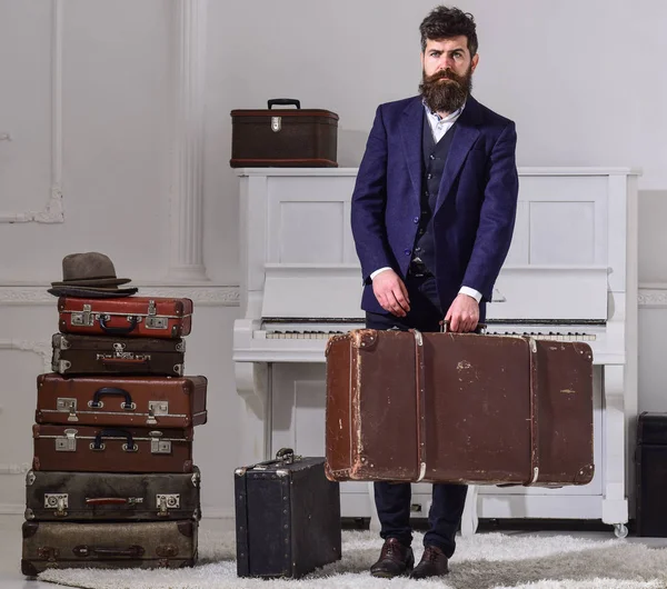 Macho elegant on strict face stands near pile of vintage suitcase, holds suitcase. Man, traveller with beard and mustache with luggage, luxury white interior background. Baggage delivery concept