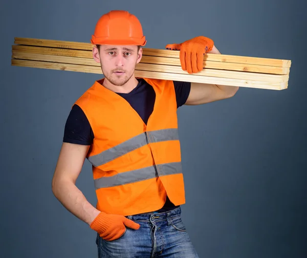 Carpintero, carpintero, obrero, constructor de cara segura lleva vigas de madera en el hombro. Concepto de trabajador resistente. Hombre en casco, sombrero duro y guantes de protección sostiene la viga de madera, fondo gris —  Fotos de Stock