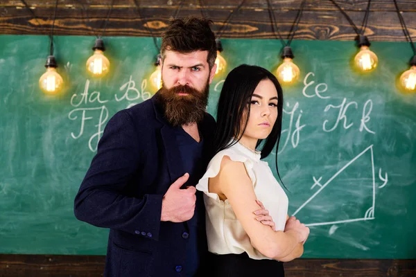 El maestro y el maestro estricto parecen confiados. Dama e hipster trabajando juntos en la escuela. Hombre con barba y sexy profesora de pie en el aula, pizarra en el fondo. Concepto de socios . — Foto de Stock