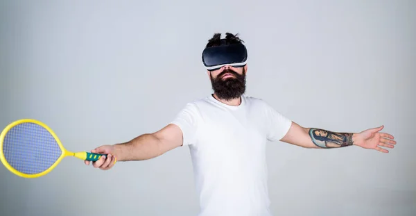 Guy with VR glasses play tennis with racket and ball. Man with beard in VR glasses beating pitch, grey background. Hipster on busy face use modern technology for sport games. Virtual sport concept