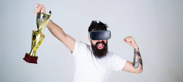 Hipster auf glücklichem Gesicht, die Faust als erfolgreiche Geste. Kerl mit kopfmontiertem Display gewann im virtuellen Spiel. Meisterkonzept. Mann mit Bart in Virtual-Reality-Brille hält Kelch, grauer Hintergrund — Stockfoto