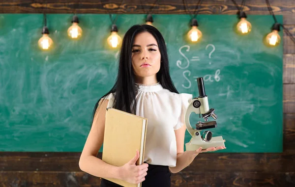 Professor de biologia detém livro e microscópio. Senhora em desgaste formal no rosto calmo na sala de aula. Lady cientista detém livro e microscópio, quadro-negro em segundo plano, espaço de cópia. Conceito de biologia — Fotografia de Stock