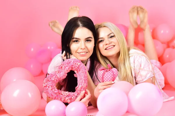 Κορίτσια βάζουν πλησίον μπαλόνια, κατέχει καρδιά παιχνίδια, ροζ φόντο. Ημέρα του Αγίου Βαλεντίνου έννοια. Ξανθιά και μελαχρινή σε χαμογελαστά πρόσωπα όνειρα σχετικά με την αγάπη και την ημερομηνία. Αδερφη, φίλοι με τις πυτζάμες στις Πιτζάμες κόμμα — Φωτογραφία Αρχείου