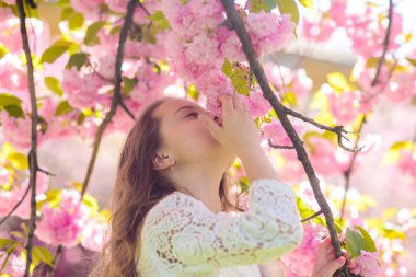 Yüz ayakta sakura çiçekler, defocused yakınındaki gülümseyerek kıza. Parfüm ve koku kavramı. Şirin çocuk bahar gününde sakura kokusu tadını çıkarın. Arka plan üzerinde uzun saçları açık, kiraz çiçeği ile kız