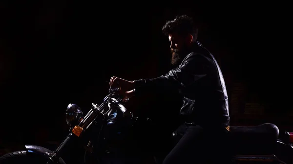 Conceito de cavaleiro nocturno. Homem com barba, motociclista de jaqueta de couro sentado em moto no escuro, fundo preto. Hipster, motociclista brutal em couro jaqueta equitação motocicleta à noite, espaço cópia — Fotografia de Stock