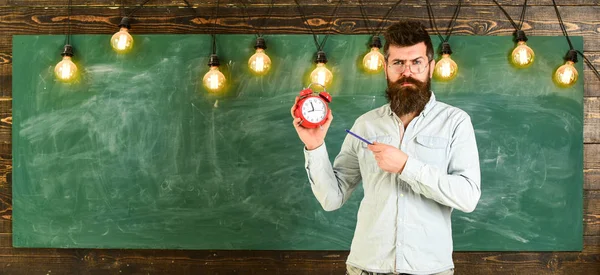 Teacher in eyeglasses holds alarm clock and pen. Bearded hipster holds clock, chalkboard on background, copy space. Man with beard and mustache on strict face stands in classroom. Discipline concept — Stock Photo, Image