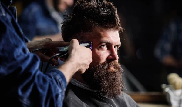 Hands of barber with hair clipper, close up. Hipster bearded client getting hairstyle. Barbershop concept. Barber works with hair clipper. Man with beard in hairdressers chair, dark background — Stock Photo, Image