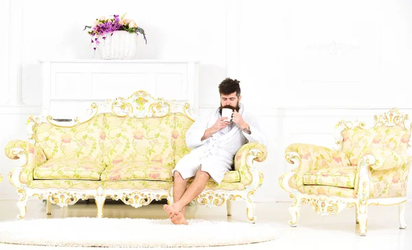 Concepto de ocio Elite. Hombre con barba y bigote disfruta de la mañana sentado en un sofá de lujo. Hombre en la cara somnolienta en albornoz, bebidas café, en hotel de lujo por la mañana, fondo blanco — Foto de Stock