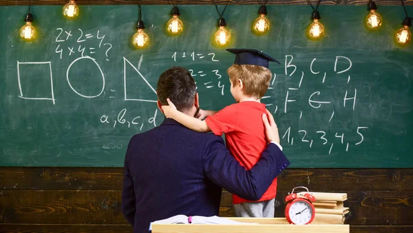 Teacher with beard, father hugs little son in classroom while discussing, chalkboard on background. Best friends concept. Child in graduate cap listening teacher, chalkboard on background, rear view — Stock Photo, Image