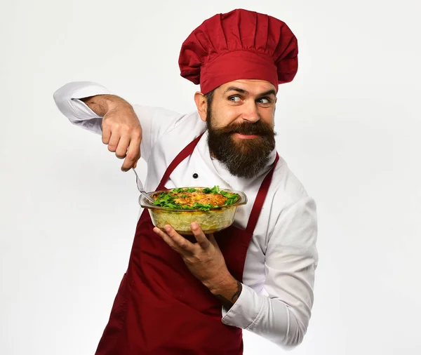 Cook with tricky face in burgundy uniform holds baked dish. — Stock Photo, Image