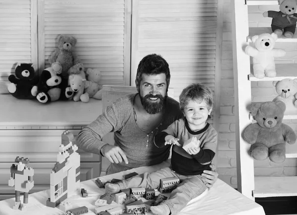 Papá y el niño construyen bloques de plástico. Hombre con barba y niño juegan cerca de juguetes y pared de madera en el fondo. Padre e hijo con caras felices hacen casa con ladrillos . —  Fotos de Stock