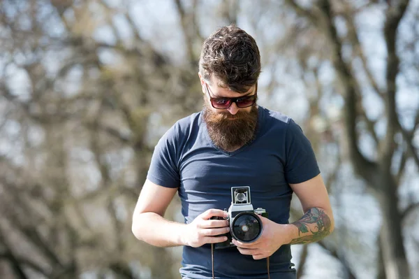 Homem barbudo trabalha com câmera vintage. Homem com barba e bigode no rosto concentrado, ramos no fundo, desfocado. Conceito de fotógrafo. Homem com barba longa ocupado com fotos de tiro — Fotografia de Stock