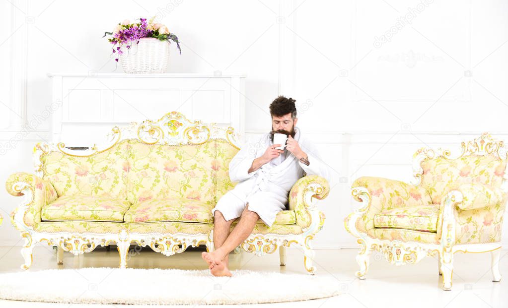 Elite leisure concept. Man with beard and mustache enjoys morning while sitting on luxury sofa. Man on sleepy face in bathrobe, drinks coffee, in luxury hotel in morning, white background