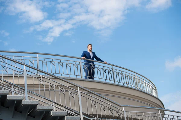 Självsäker affärsman står på balkongen på molnig blå himmel. Man i högkvalitativt slitage utomhus. Ser mot framtiden. Funderar på nya möjligheter. Förtroende och karisma. En manlig blick. Bästa managern någonsin. — Stockfoto