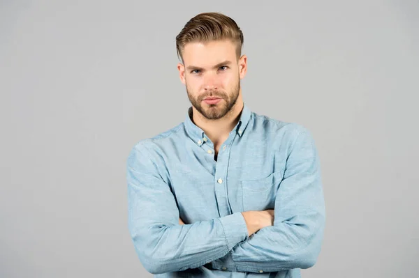 Bearded man keep arms crossed on grey background. Macho in blue tshirt with serious face. Tired after everyday work. Office dress code. Casual in style. Confidence and charisma. Young and handsome.