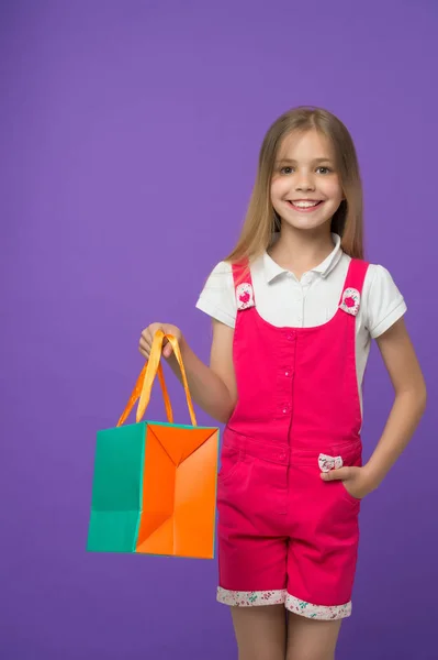 Glad tjej med shoppingväska på lila bakgrund. Lite shopaholic leende med papperspåse. Barn shopper i mode overall. Semester förberedelser och firande. Barndomen. Shopping och försäljning — Stockfoto
