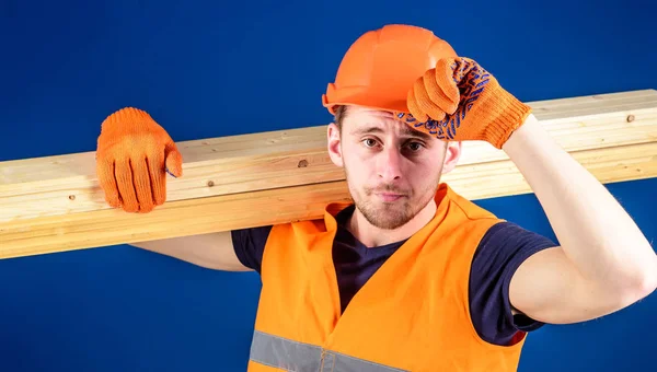 Woodworker concept. Man in protective gloves holds visor of helmet, corrects hard hat on head, blue background. Carpenter, woodworker, labourer, builder carries wooden beam on shoulder — Stock Photo, Image