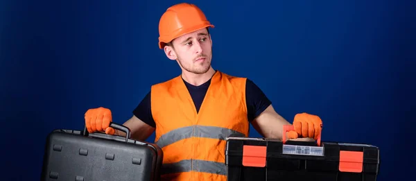 Toolbox and equipment concept. Worker, repairer, repairman, builder on thoughtful face choosing equipment for work. Man in helmet, hard hat holds toolbox and suitcase with tools, blue background — Stock Photo, Image