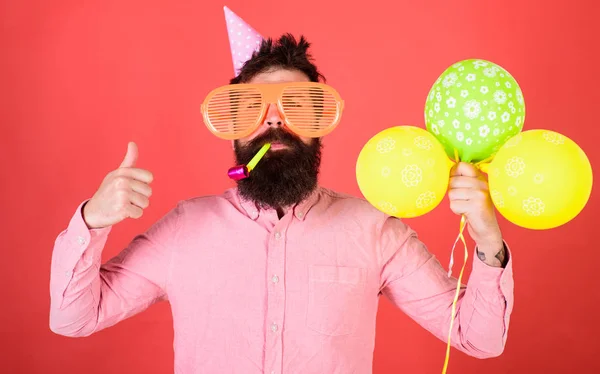 Surprise birthday party with balloons decoration. Bearded man in pink shirt showing thumb up. Hipster with huge glasses and party whistle isolated on red background. Man organizing birthday for kids
