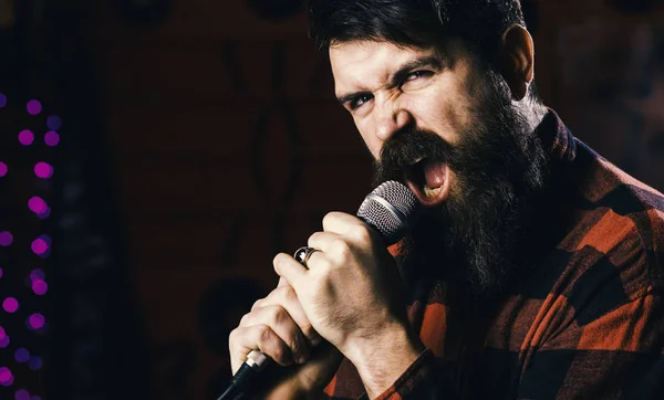 Guy in checkered shirt on black background, defocused. Young man singing with microphone. Hipster with beard