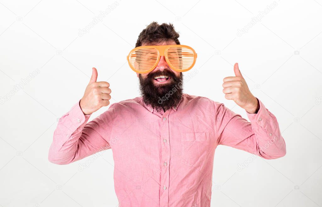 Cheerful mood concept. Hipster looking through of giant striped sunglasses. Guy with beard shows thumbs up. Man with beard and mustache on happy face wears funny big eyeglasses, white background