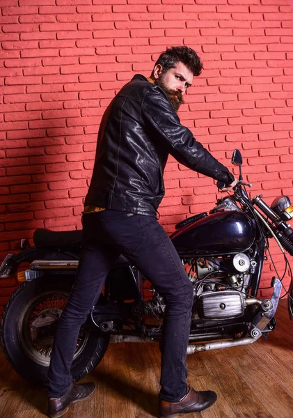 Man with beard, biker in leather jacket near motor bike in garage, brick wall background. Hipster, brutal biker on serious face in leather jacket sits down on motorcycle. Masculine hobby concept