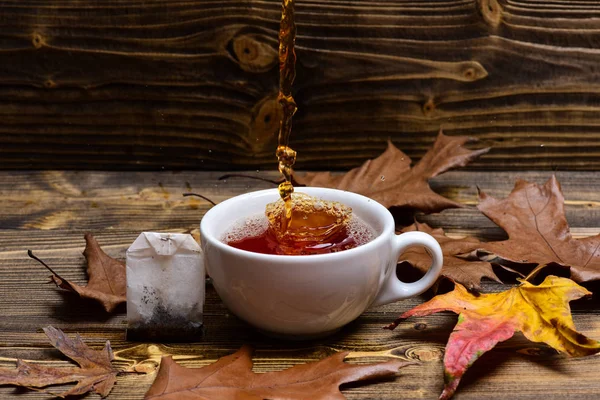 Brewing tea concept. Cup pouring with water or tea with splashes on dark wooden background. Cup, autumn leaves and teabag on wooden table. Mug pouring with liquid with splashes and drops of water