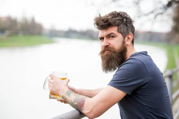 Man with long beard looks relaxed. Man with beard and mustache on calm face, river background, defocused. Bearded man holds beer mug, drinks beer outdoor. Craft beer concept