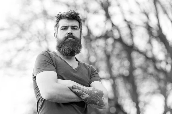 Brutal bearded man with gun and roses tattoo standing with his arms crossed at his chest. Handsome man with blue eyes and cool hairstyle posing outdoors