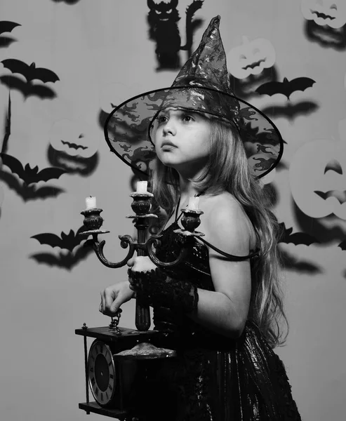 Halloween festa e decorações conceito. Menina com o rosto interessado no fundo rosa com decoração assustadora. Kid em fantasias de bruxas assustadoras segura relógio velho e lustre — Fotografia de Stock