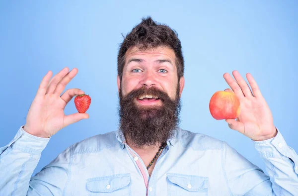 Beneficios de verano. Hombre barbudo sonriente sostiene manzana y fresa en las manos fondo azul. La fruta y la baya en las manos la alternativa sana. Concepto nutricional de la vitamina fruta. Vitamina para dietas sanitarias — Foto de Stock