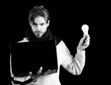 Beyaz süveterli, siyah arka planda örülmüş sakallı yakışıklı bir adam ya da sakallı bir adam dizüstü bilgisayar, lamba ya da ampul tutuyor.