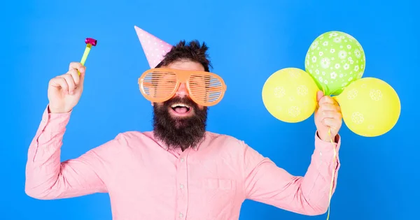 Человек с бородой и усами на счастливом лице держит воздушные шары, синий фон. Хипстер в огромных очках празднует день рождения. Концепция вечеринки. Парень в шляпе с атрибутами праздника празднует — стоковое фото