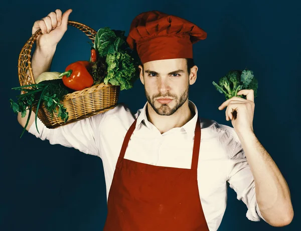 Chef in burgundy uniform with broccoli and other vegetables. Man with beard on green background. Vegetarian cooking concept.