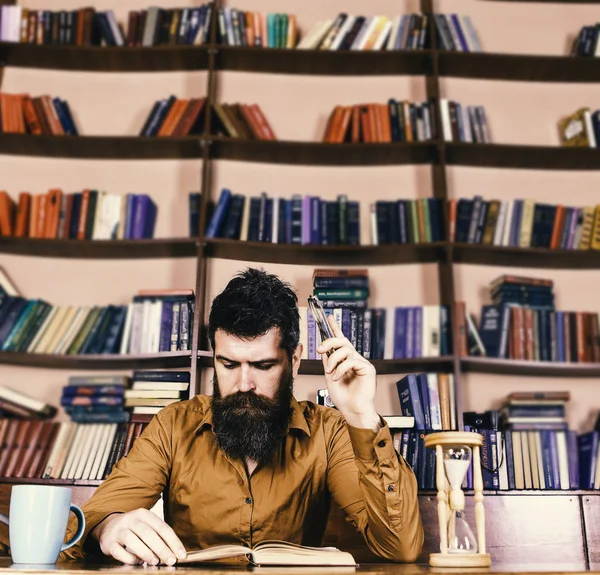 Profesor o estudiante con barba estudiando en la biblioteca. Hombre ocupado leyendo libros, estanterías en el fondo. Concepto de educación y ciencia. Científico leyendo, se sienta a la mesa con taza y reloj de arena —  Fotos de Stock