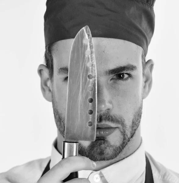 Человек в шапке и фартуке держит острый клинок у лица. Повар работает на кухне. Кухонные принадлежности и кулинарная концепция . — стоковое фото