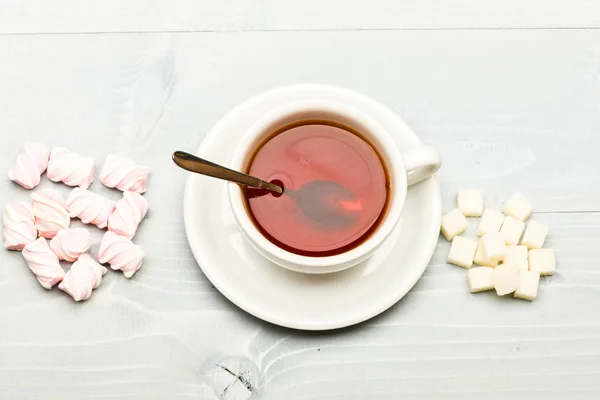 Te paus koncept. Mugg med svart te och godis. Mugg fylld med svart bryggt te, sked och högen av marshmallow och raffinerat socker på vit trä bakgrund. Dryck och godis beredd som dessert — Stockfoto